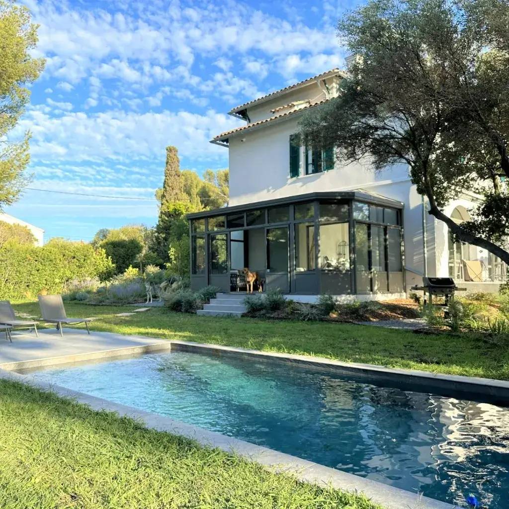 Cette image montre une maison spacieuse avec une véranda moderne, entourée de verdure, avec une piscine en premier plan sous un ciel bleu