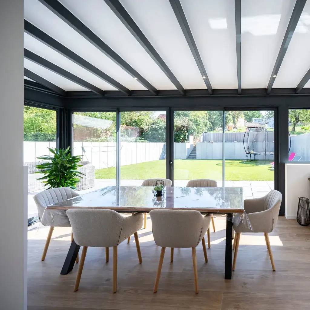 Cette véranda lumineuse avec un toit en poutres apparentes offre une vue dégagée sur un jardin verdoyant, idéale pour des repas convivia