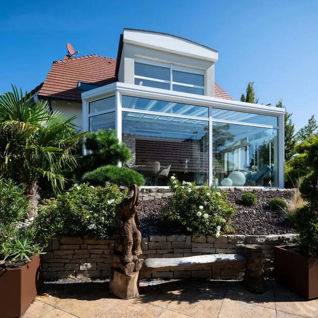 Une élégante véranda moderne avec de grandes baies vitrées s'ouvrant sur un jardin soigneusement aménagé sous un ciel bleu clair.