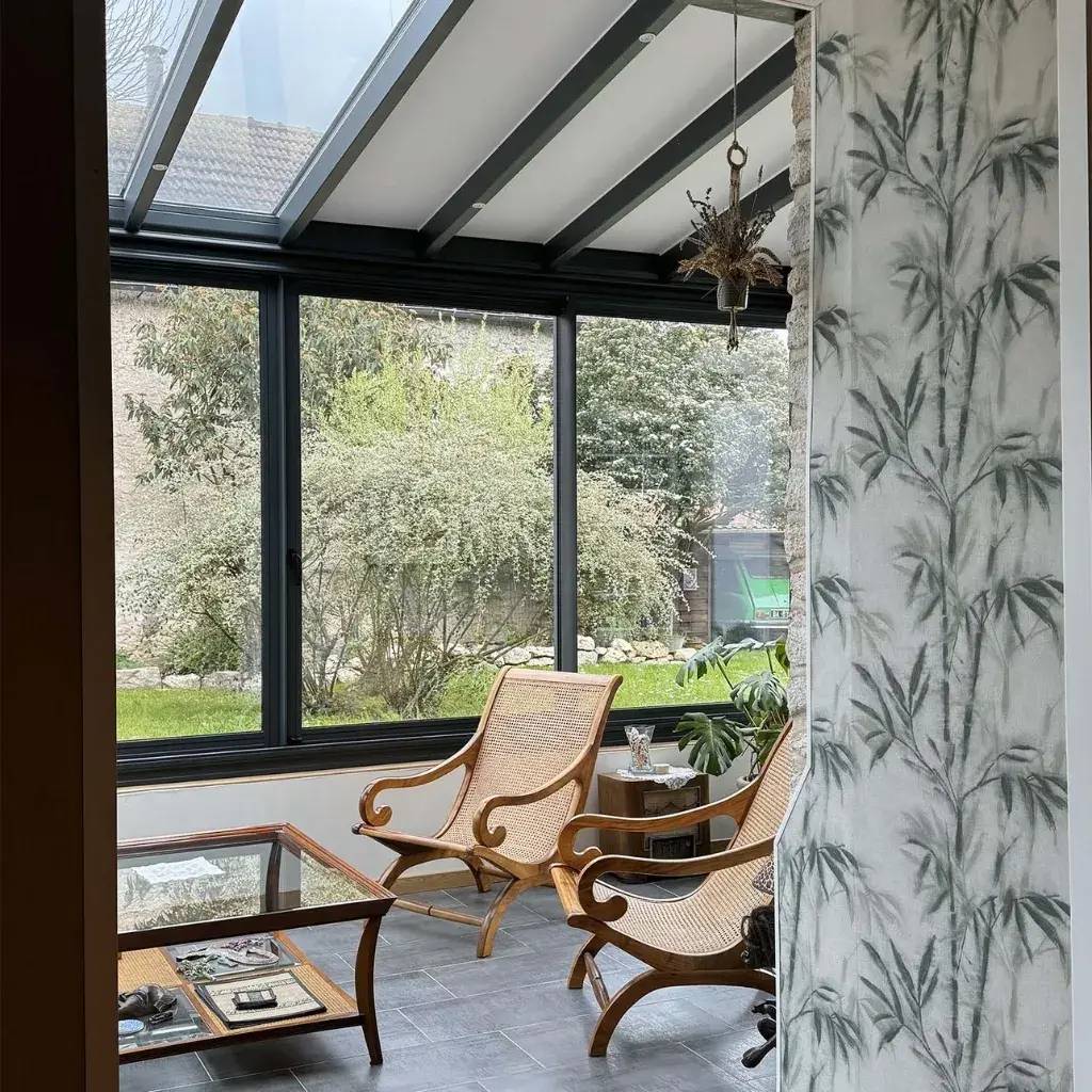 Une véranda lumineuse avec de grandes fenêtres, des fauteuils en osier et une table basse en verre, offrant une vue sur un jardin verd