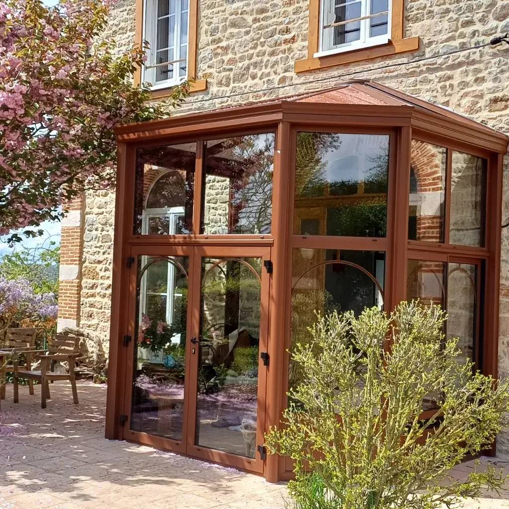 Une belle véranda en bois et verre adossée à une maison en pierre, entourée de végétation printanière.
