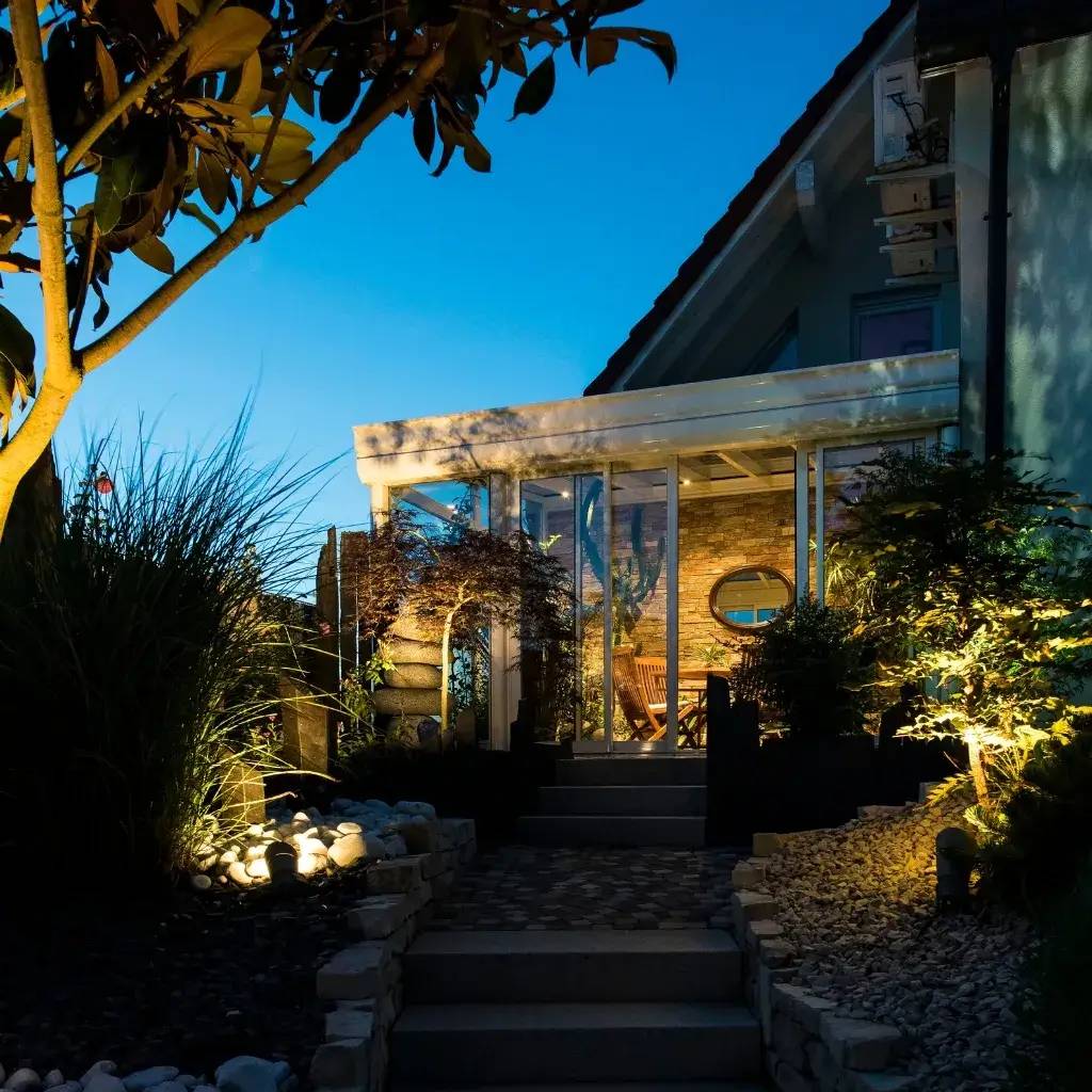 Une véranda lumineuse en soirée entourée d'un jardin magnifiquement aménagé avec des éclairages subtils.