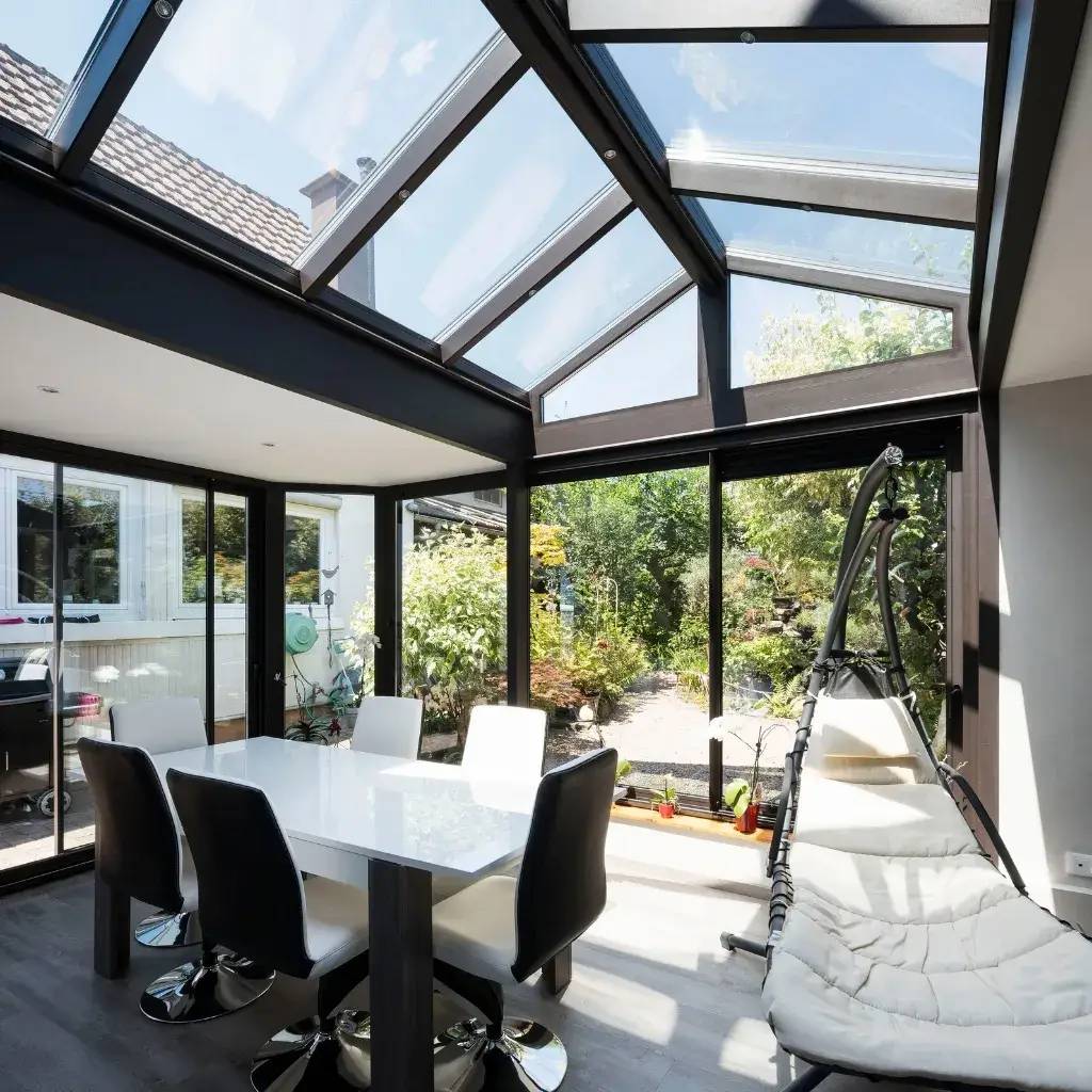 Une véranda moderne avec un toit en verre transparent inonde une salle à manger élégante et lumineuse de lumière naturelle, offrant une vue sur un