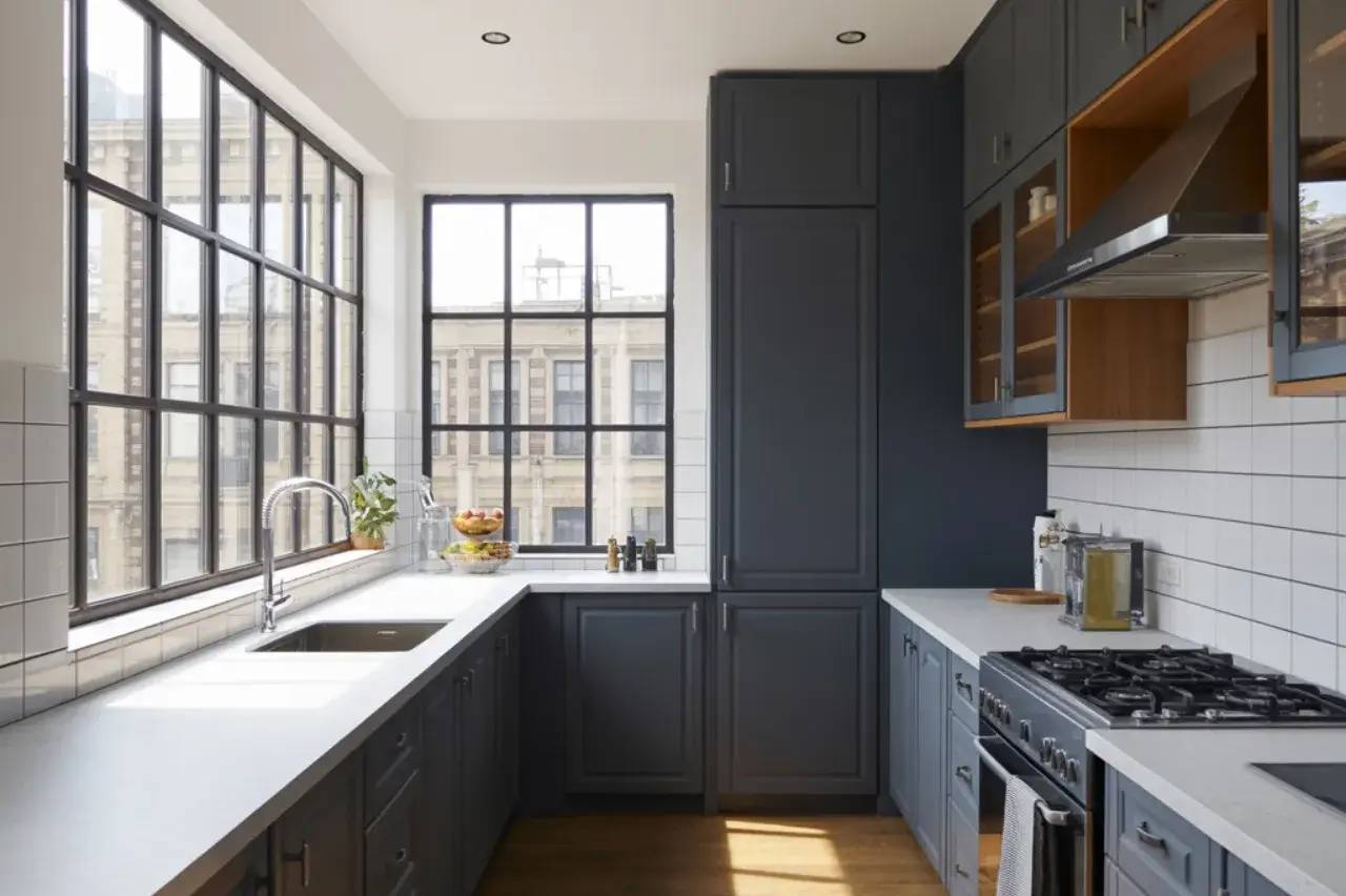 Cuisine moderne et lumineuse avec de grandes fenêtres, des armoires sombres et un plan de travail clair.