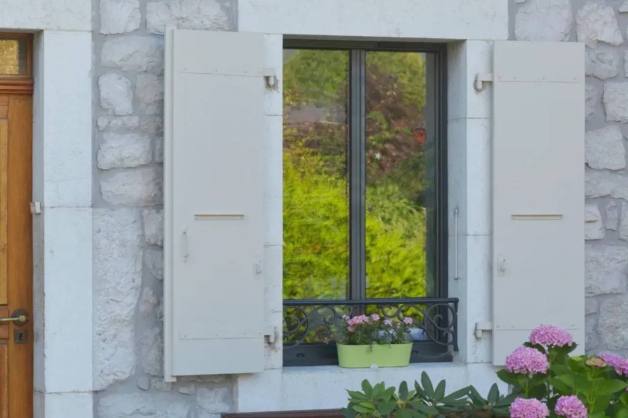 Une fenêtre à volets blancs est encadrée par un mur en pierre, ornée d'une jardinière de fleurs roses.
