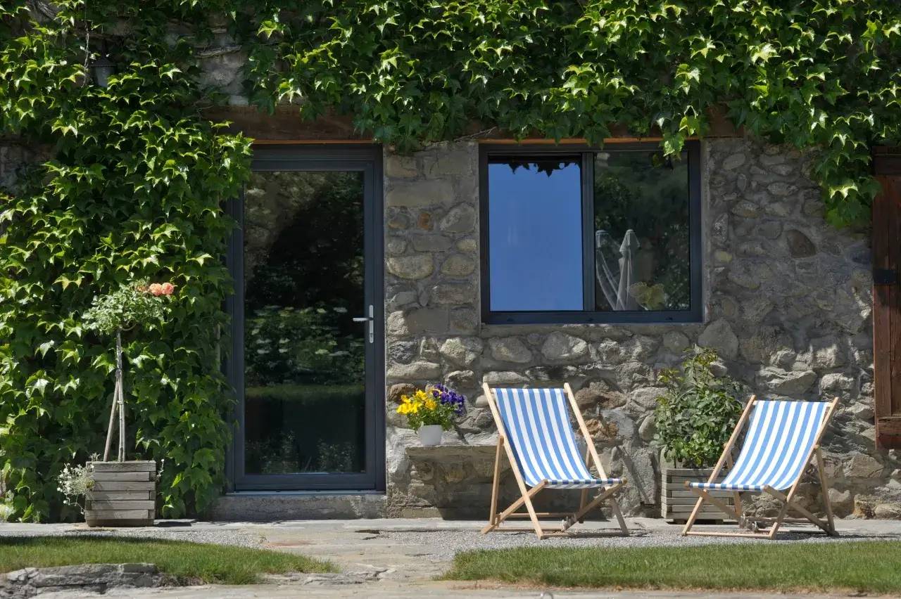 Façade en pierre d'une maison avec des chaises longues rayées bleu et blanc entourées de verdure grimpante.
