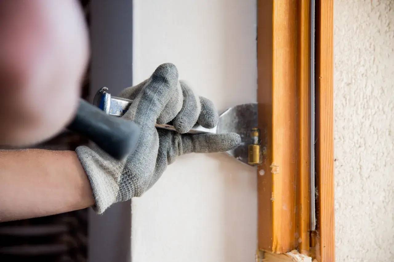 Une personne en gants utilise un outil pour travailler sur une charnière de fenêtre en bois.