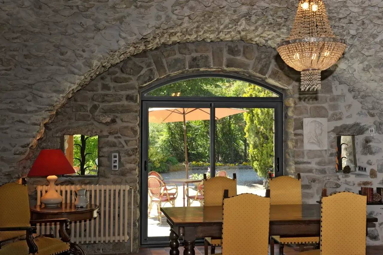 Une élégante salle à manger en vieilles pierres avec un lustre en cristal, donnant sur une terrasse ensoleillée entourée de verdure.