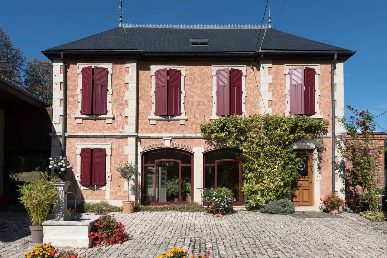 Une maison en pierre à deux étages avec des volets bordeaux, entourée de végétation et de fleurs colorées, sous un ciel bleu