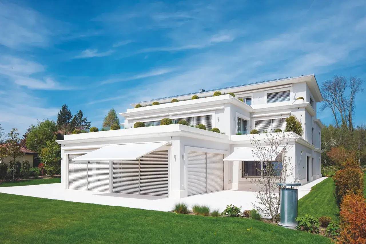 Une élégante maison contemporaine blanche avec de grandes fenêtres, entourée d'un jardin verdoyant sous un ciel bleu.