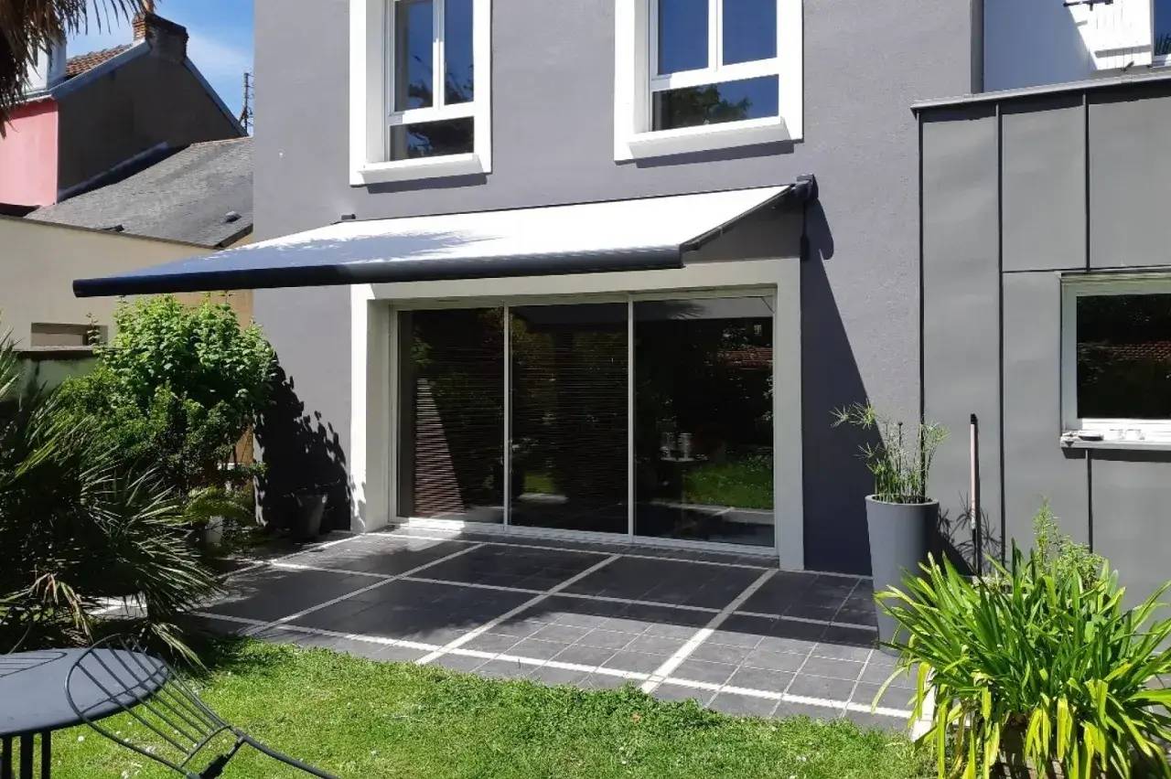 Une terrasse en dalles grises est ombragée par un store sur une maison moderne avec un jardin vert et des plantes en pot.