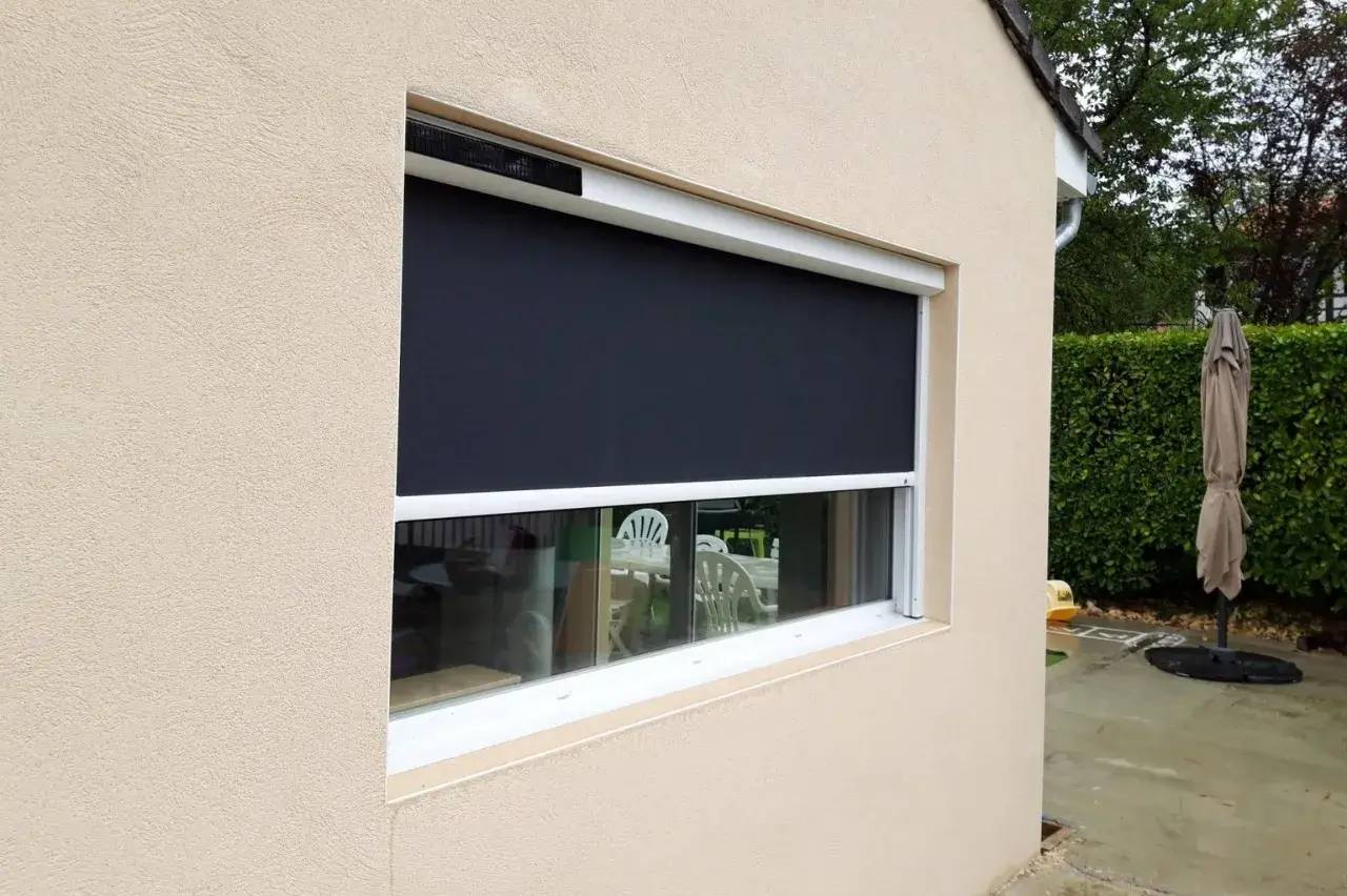 Une fenêtre à moitié ouverte avec un store occultant extérieur gris sur une façade beige.