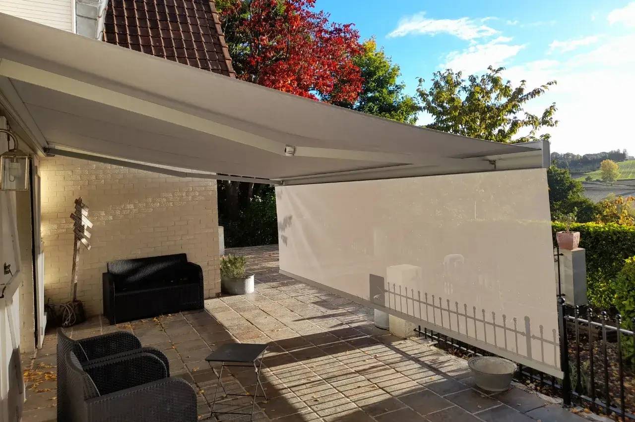 Une terrasse accueillante avec des fauteuils en rotin noir, sous un grand store, entourée de verdure et baignée de lumière naturelle.
