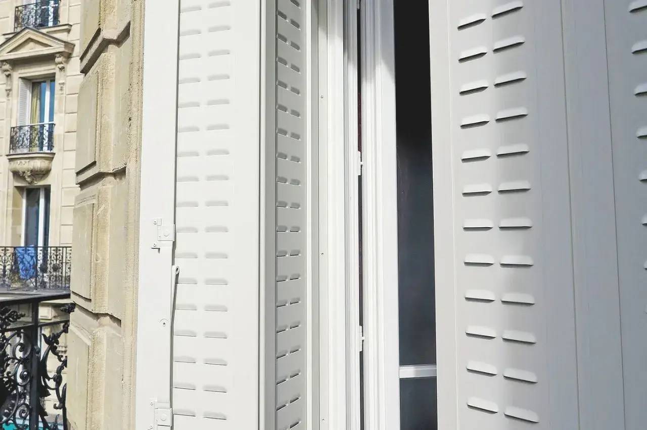 Vue sur un balcon parisien avec des volets blancs ouverts laissant entrer la lumière.