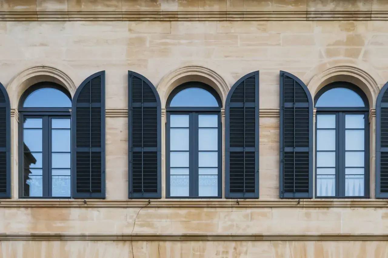 Façade d'un bâtiment classique avec des fenêtres arquées et des volets noirs contrastant sur un mur de pierre beige.