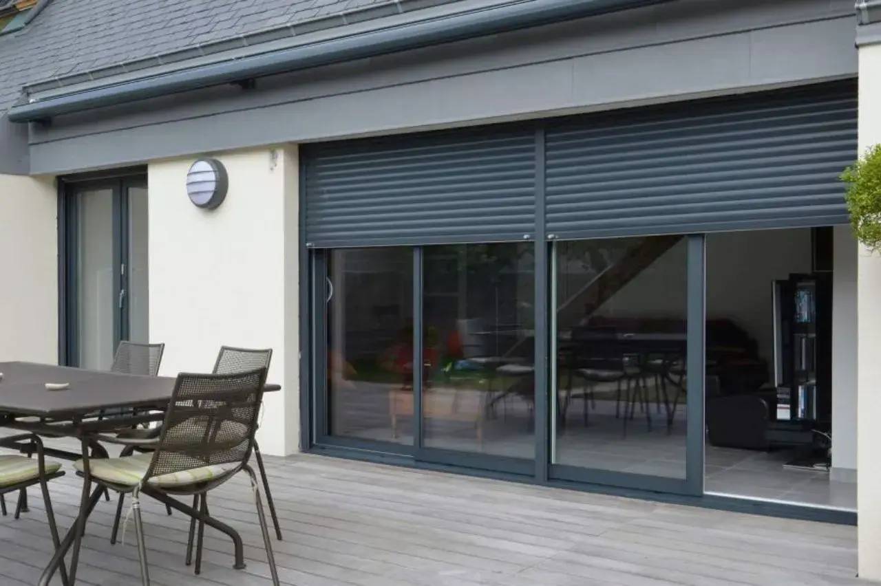 Une terrasse moderne avec des chaises et une table en métal, adossée à une maison équipée de volets roulants gris sur de grandes ba