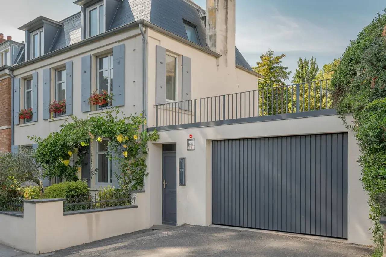 Maison de style moderne avec façade crépie, volets gris, garage intégré et jardin fleuri.