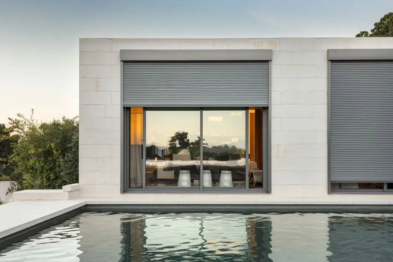 Vue extérieure d'une maison moderne avec des volets roulants fermés et une piscine à l'avant, reflétant le paysage environnant.
