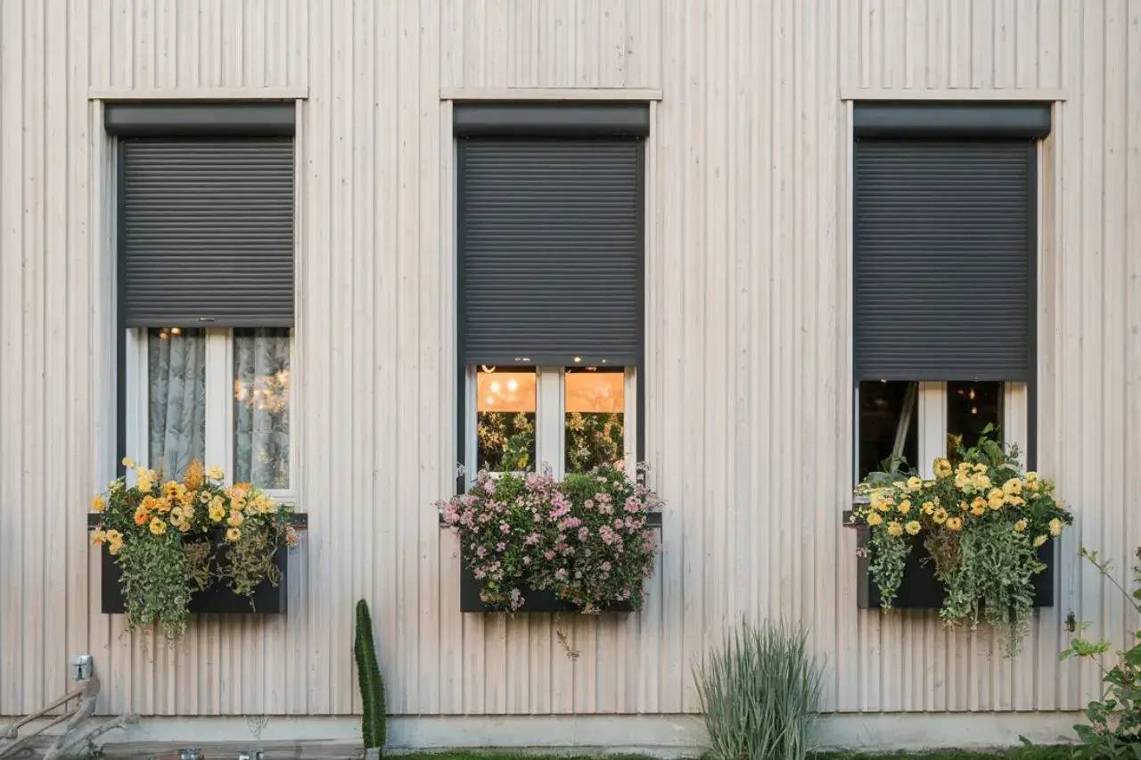 Trois fenêtres ornées de volets roulants gris foncé et de jardinières fleuries décorent la façade en bois clair d'une maison
