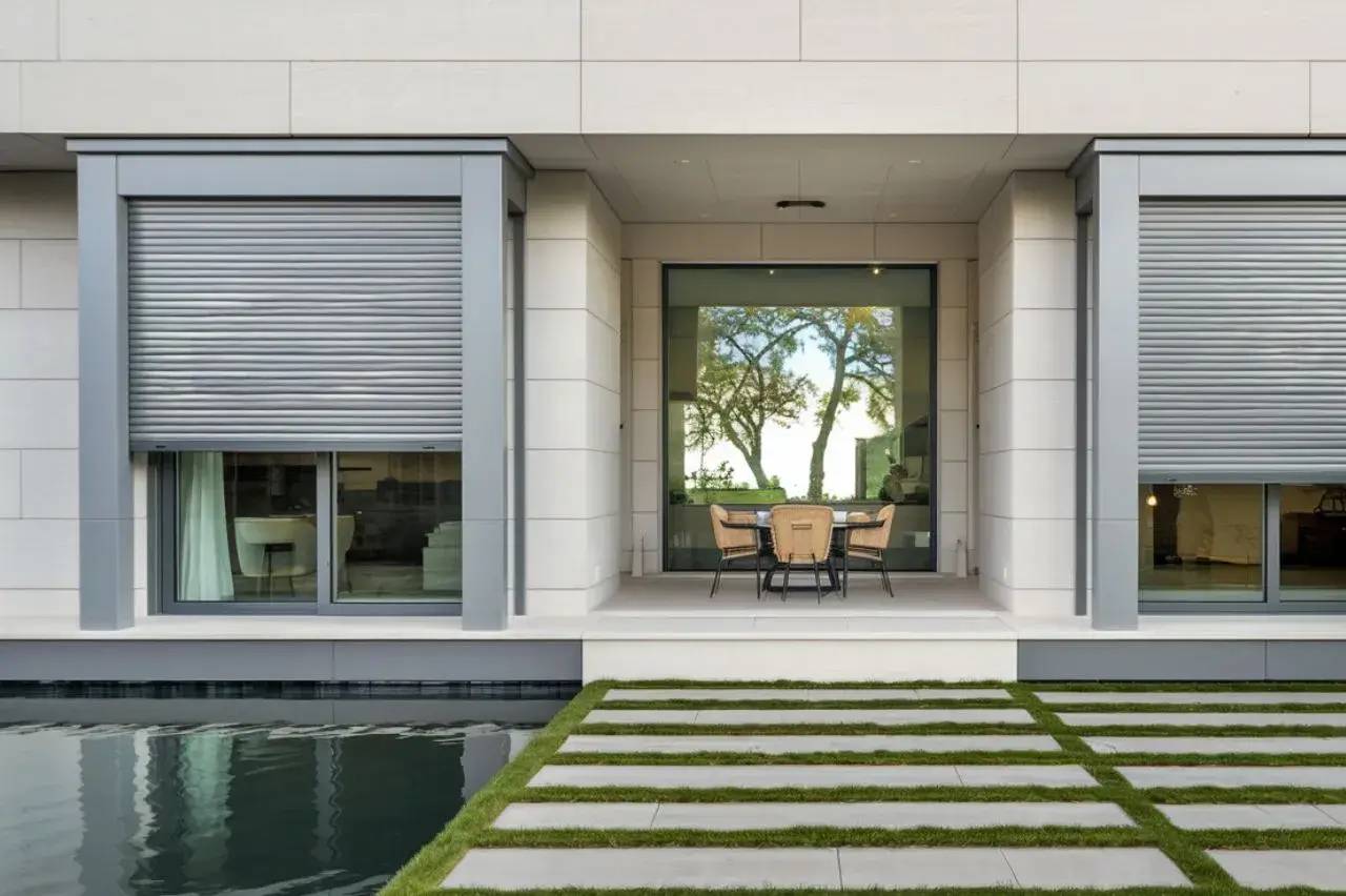 Façade moderne d'une maison avec un patio élégant, des stores métalliques, et une vue sur un jardin avec une piscine.