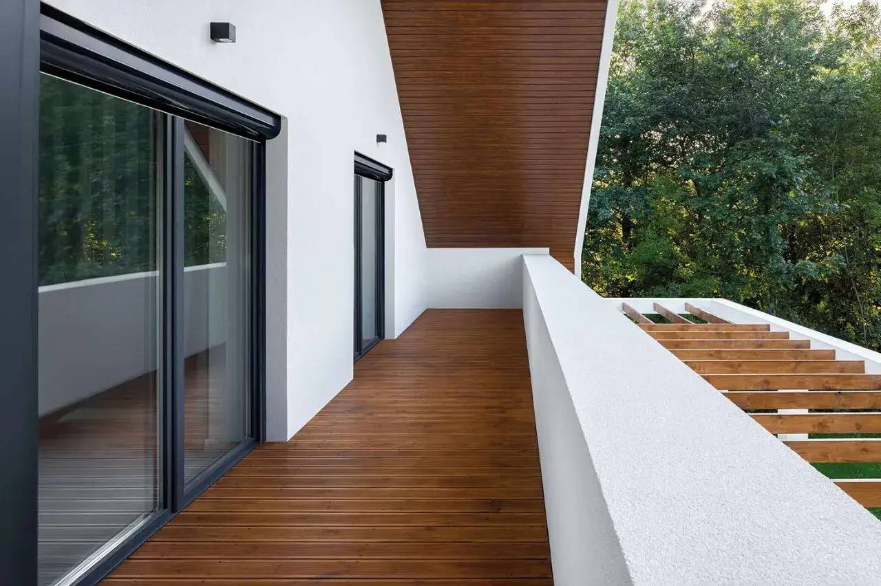 Une terrasse moderne en bois avec une balustrade blanche et de grandes baies vitrées, offrant une vue sur la nature environnante.