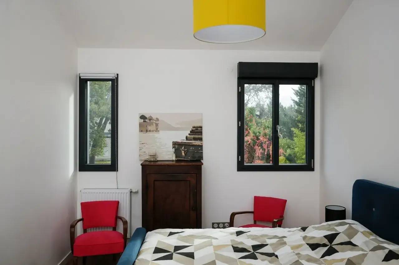 Une chambre lumineuse avec des touches de couleurs vives, comprenant deux fenêtres, des fauteuils rouges, et un luminaire jaune au-dessus d'un