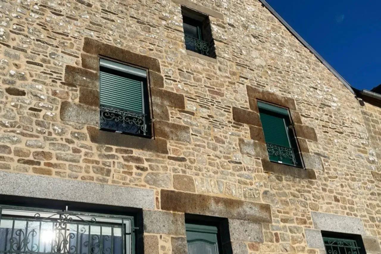 Façade d'une maison en pierres avec des volets roulants verts et encadrements en pierre sombre.