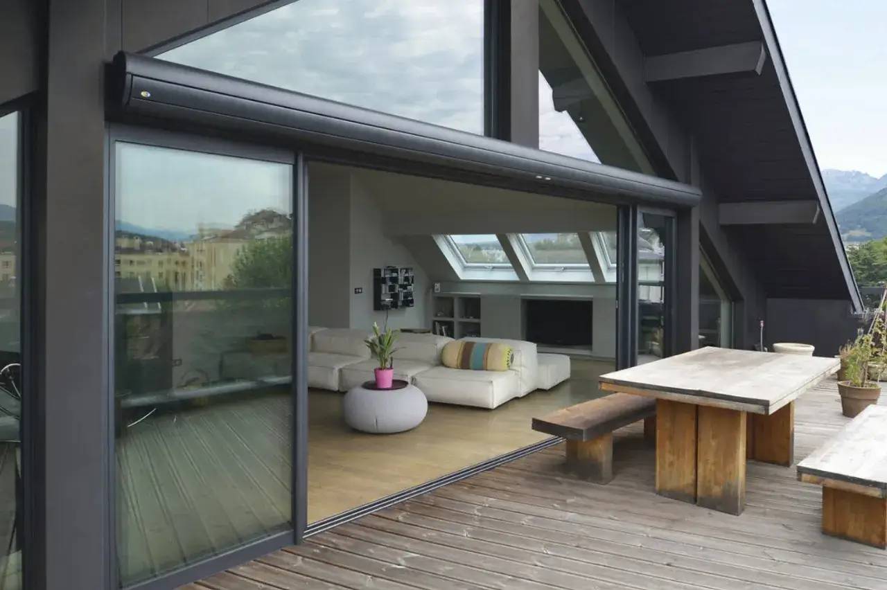 Une terrasse en bois spacieuse avec une table en bois rustique connectée à un salon intérieur moderne et lumineux à travers de grandes baies vitr