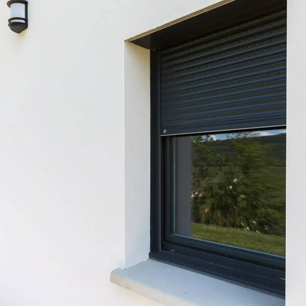 Une fenêtre moderne avec des volets roulants métalliques noirs, sur une façade blanche, offrant une vue partielle sur un paysage verdoyant.