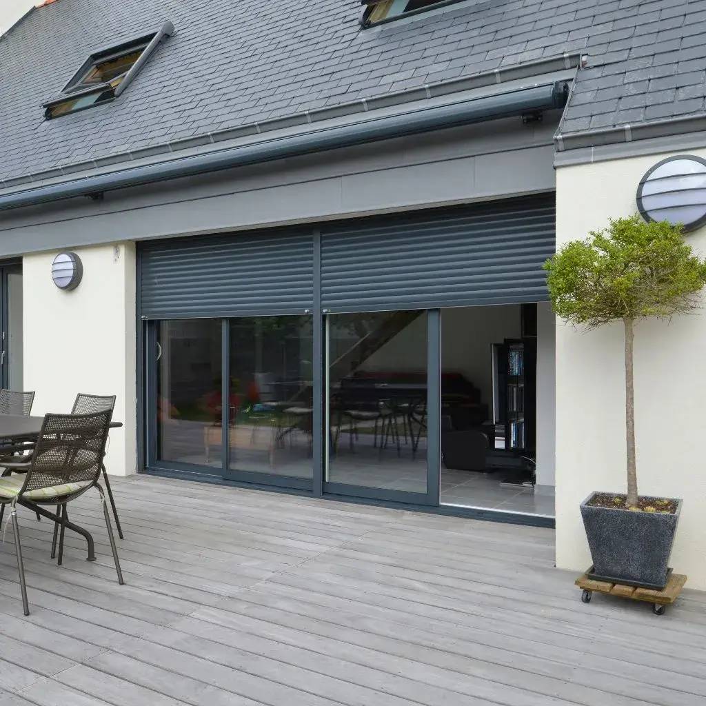 Une terrasse moderne en bois avec des chaises en métal et de grandes fenêtres coulissantes dotées de volets roulants.