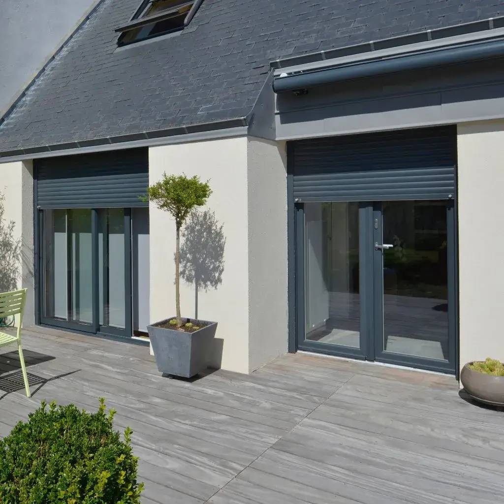 Maison moderne avec façade en crépi et volets roulants, donnant sur une terrasse en bois avec des plantes en pots.