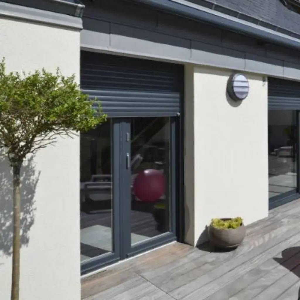 Une terrasse moderne avec sol en bois, une plante en pot, une grande baie vitrée et un store métallique.