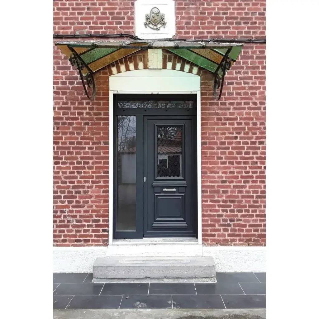 Une porte d'entrée en bois sombre est encadrée dans un mur de briques rouges, surmontée d'un auvent en verre et fer