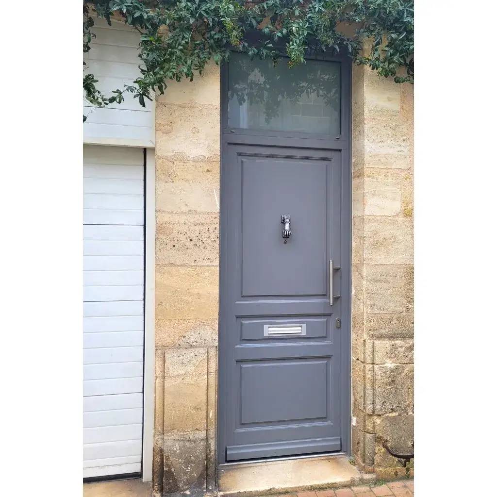 Cette image montre une porte d'entrée grise élégante avec une poignée moderne et une boîte aux lettres, encadrée par un mur en pierre claire