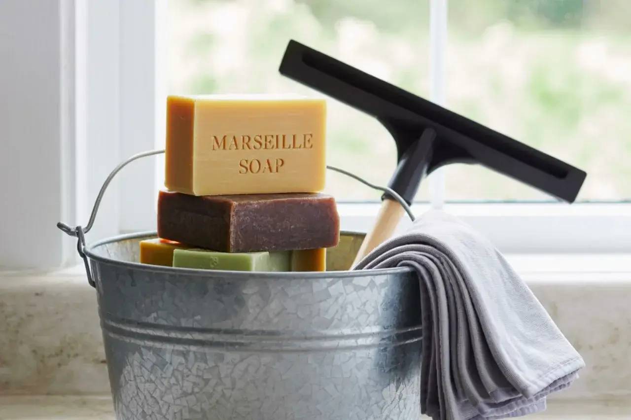 Un seau en métal contient des savons de Marseille empilés, un racloir et une serviette grise, posé devant une fenêtre lumineuse