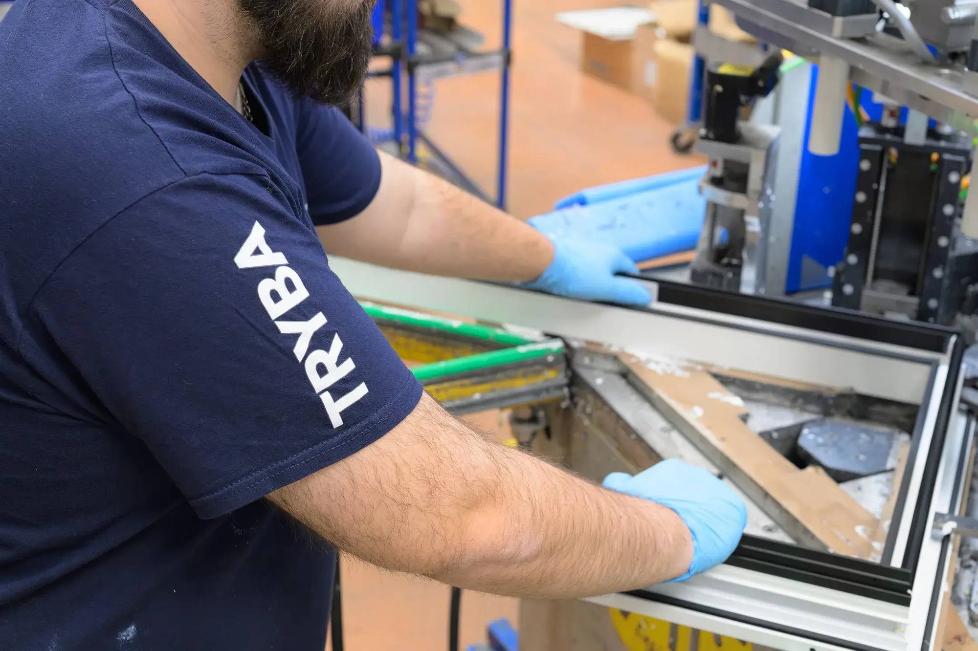 Un ouvrier portant des gants et un t-shirt bleu assemble un cadre de fenêtre dans une usine.