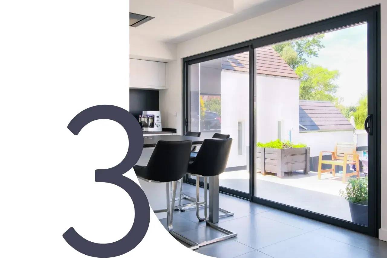 Une cuisine moderne avec des chaises hautes, baignée de lumière naturelle grâce à de grandes baies vitrées donnant sur une terrasse.