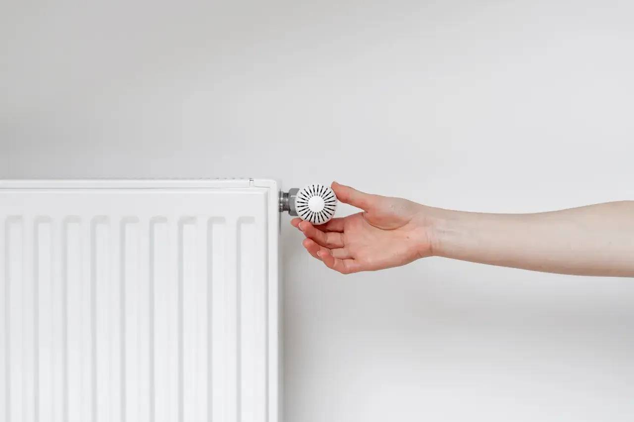 Une main ajuste le bouton d'un radiateur blanc sur un fond mural uni.