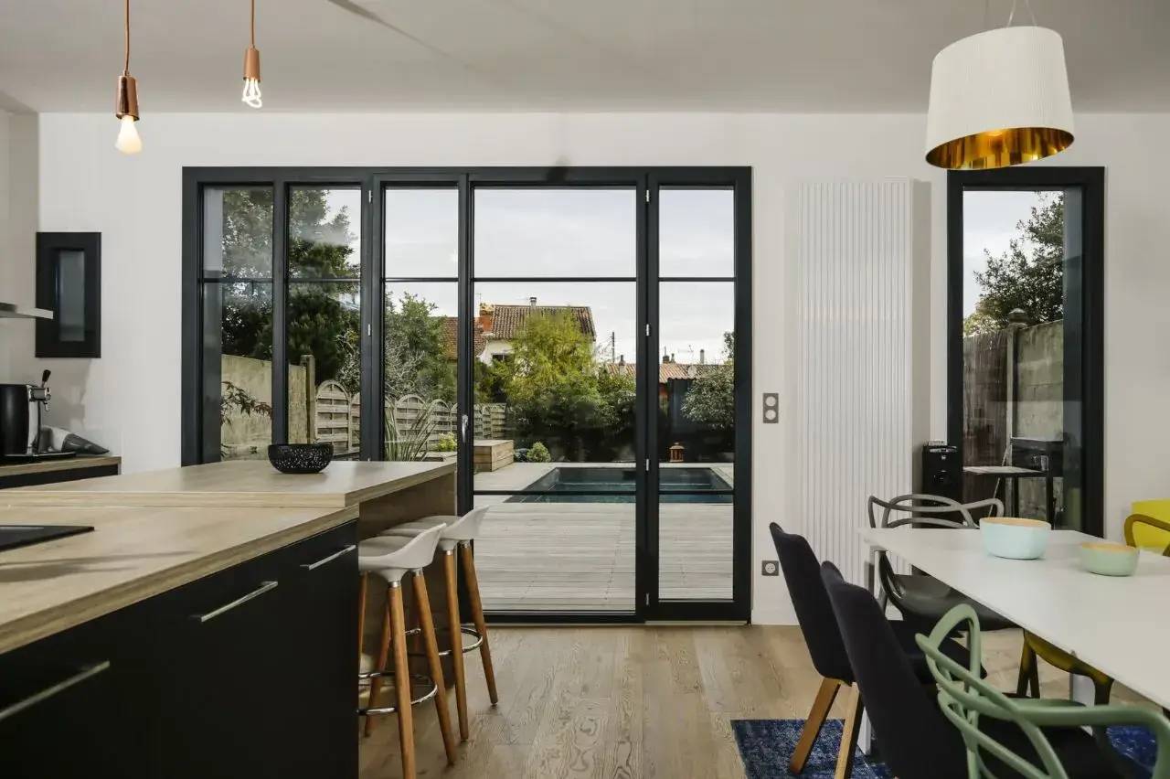 Une cuisine moderne ouverte sur une terrasse avec piscine, mêlant éléments en bois et touches de noir, illuminée par une grande baie vitrée.