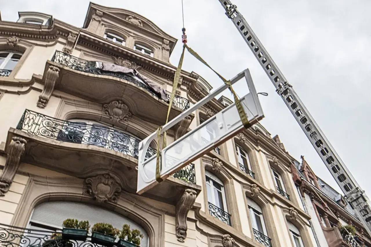 Une grue soulève une structure métallique devant un bâtiment à l'architecture noble, ornée de balcons en fer forgé.