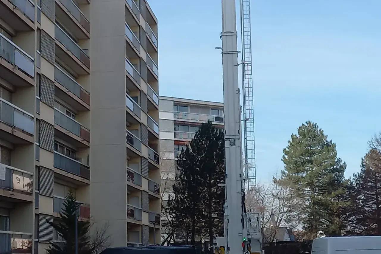 Une grue blanche est installée devant des immeubles résidentiels, avec des arbres en arrière-plan.