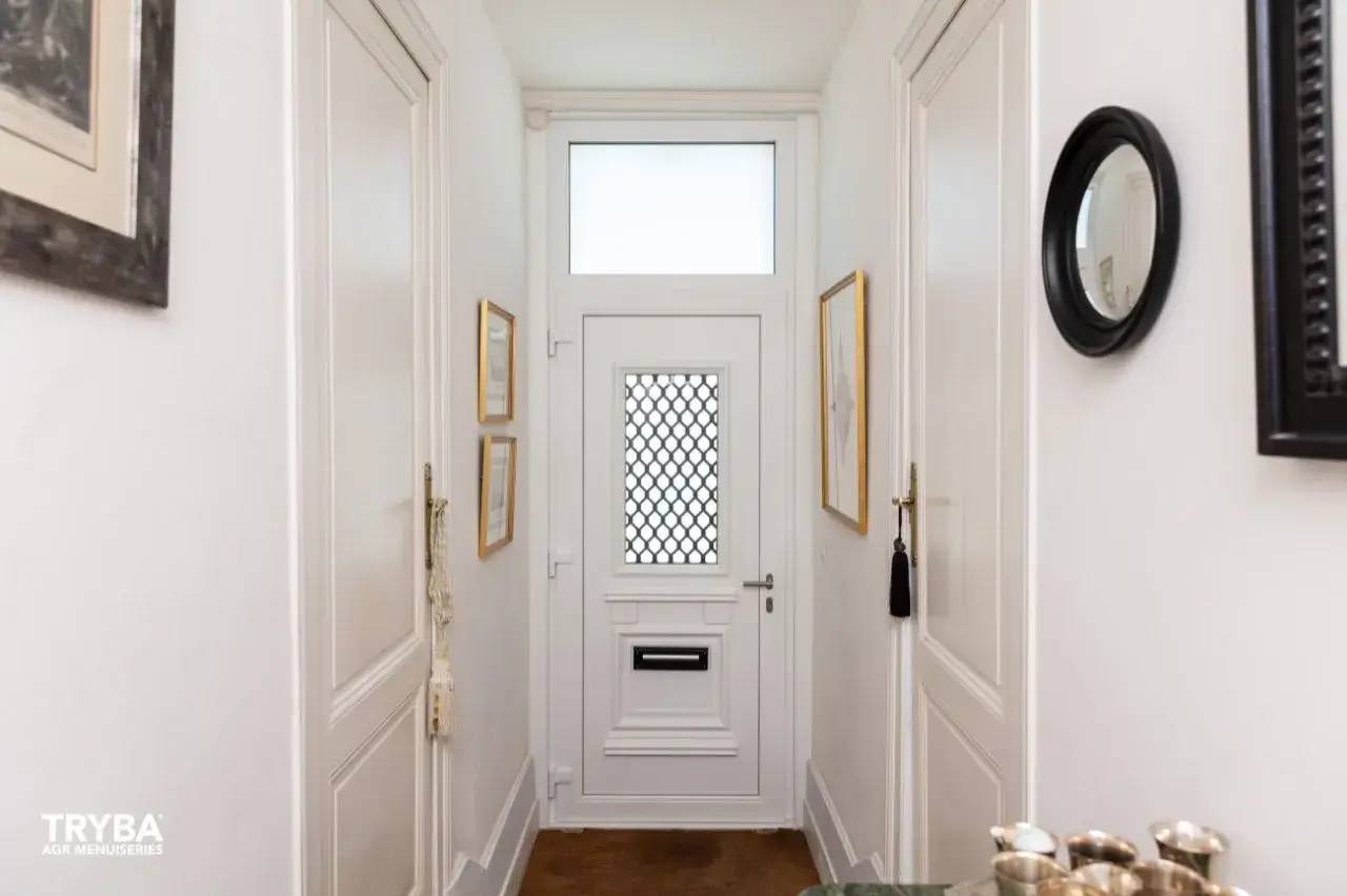 Un couloir lumineux au décor épuré et élégant, avec une porte blanche à motifs géométriques et des cadres dorés accrochés aux murs
