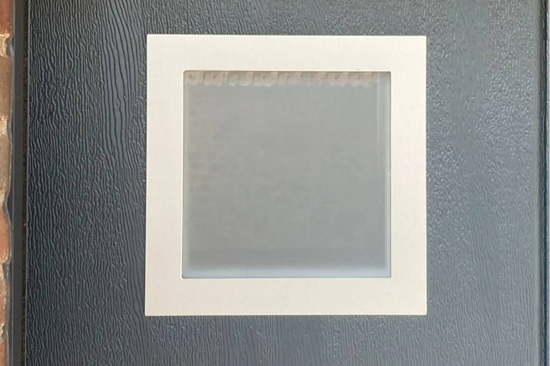 Une fenêtre carré avec un cadre blanc est fixée sur une porte grise texturée.