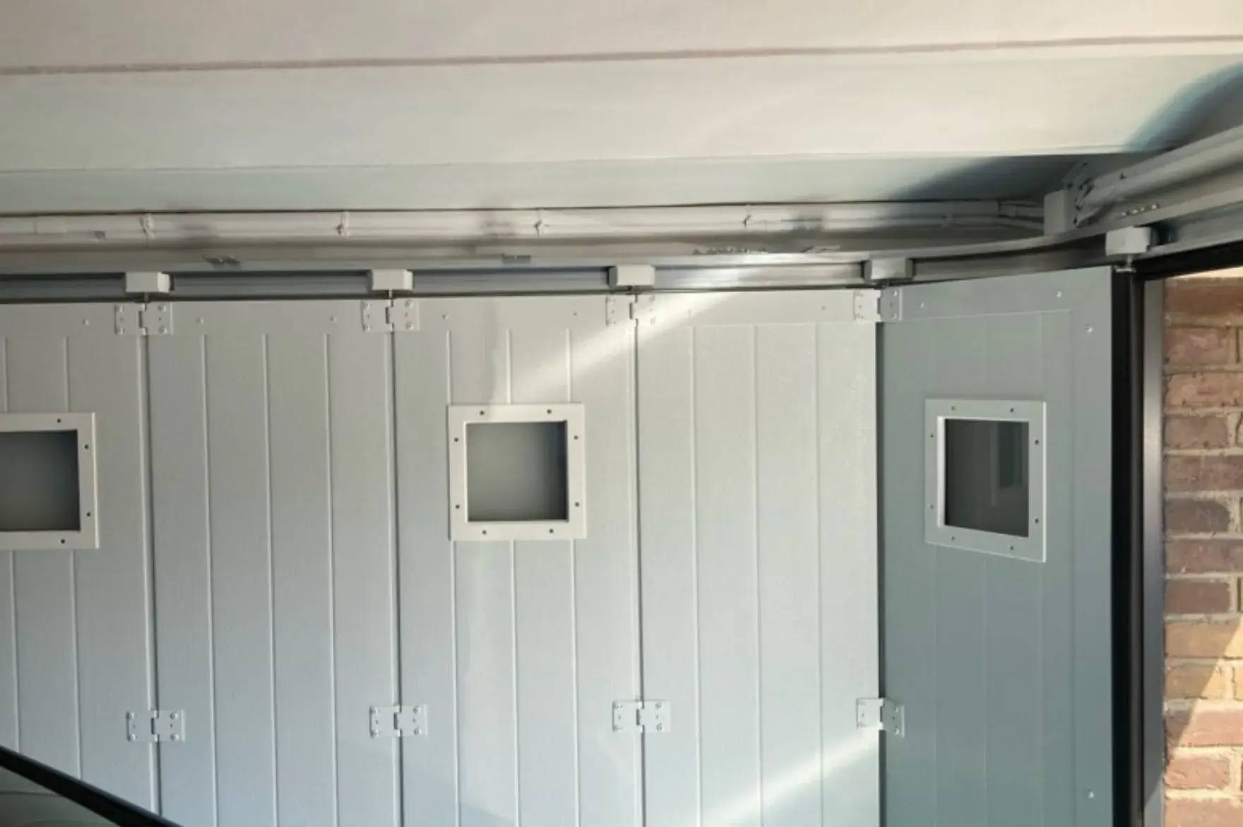 L'image montre l'intérieur d'une porte de garage en métal blanc, avec deux petites fenêtres rectangulaires, sur fond de murs en briques
