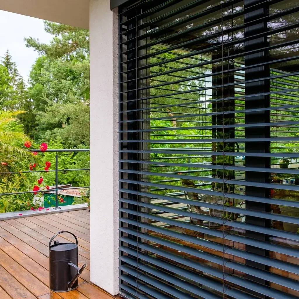 Une terrasse en bois avec un arrosoir noir et une vue sur un jardin verdoyant, bordée par de grandes baies vitrées équipées