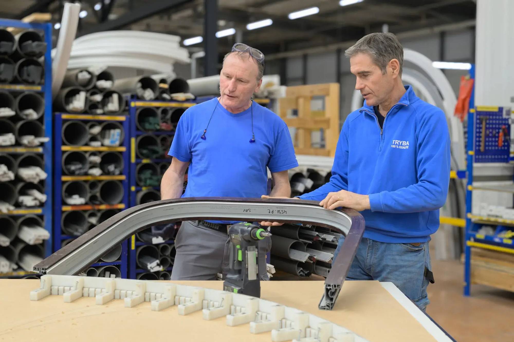 Deux hommes en bleu inspectent un cadre de fenêtre incurvé dans un atelier industriel.