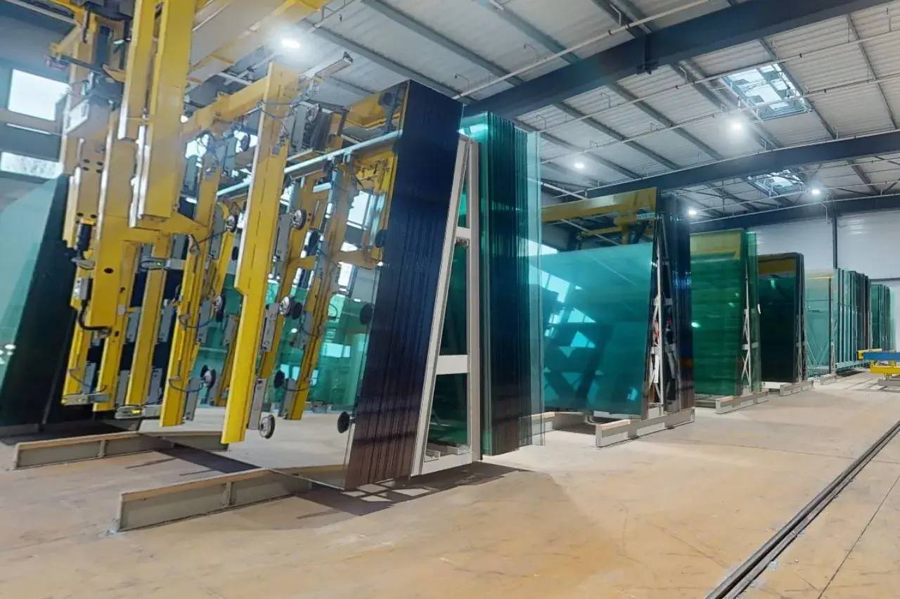 Un entrepôt industriel avec de grandes vitres en verre turquoise empilées sur des supports métalliques et des structures jaunes de manutention.